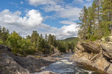 Fototapeta na wymiar River in Norwegian landscape with rocks trees and blue sky with white clouds. There is sunshine and shadows. View up stream.