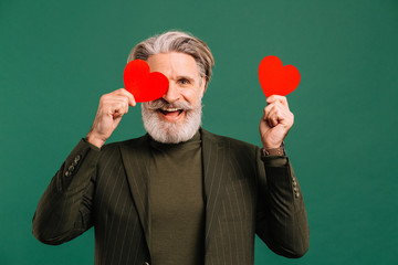 Happy bearded and mustache mature man in khaki suit holding two red hearts near eyes greeting...