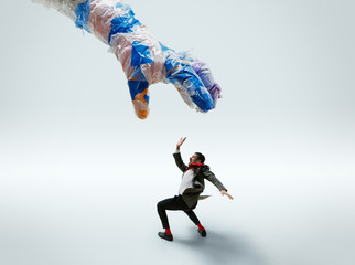 Young caucasian man avoiding big plastic hand on white studio background. Male model in office attire fighting in motion and action. Looks angry, scared, pressured. Eco problems, pollution, recycling.