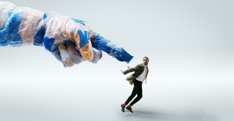 Young caucasian man avoiding big plastic hand on white studio background. Male model in office attire fighting in motion and action. Looks angry, scared, pressured. Eco problems, pollution, recycling.