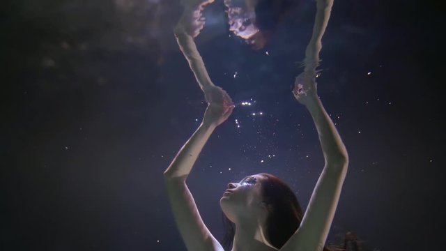 Close-up portrait of a face of a dark-haired girl with rhinestones on her face, who is underwater on a blue background, she is reflected from the surface of the water, pulling her hands up.