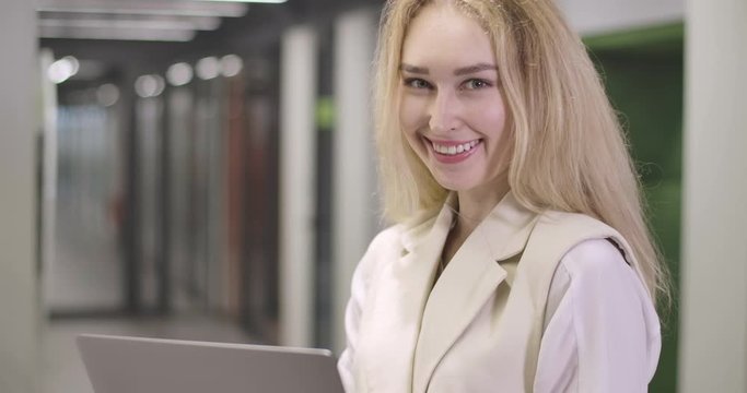 Portrait of beautiful blond Caucasian woman standing with laptop in open space office, looking at camera and smiling. Successful young businesswoman posing at workplace. Cinema 4k ProRes HQ.