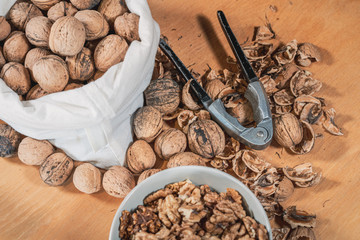 Close up view of whole walnuts in a cotton bag, a nutcracker and scattered nuts and shells on a wooden table. Walnut kernels in a bowl