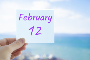 February 12th. Hand holding sticker with text February 12 on the blurred background of the sea and sky. Copy space for text. Month in calendar concept