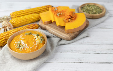 pumpkin cream soup in a bowl on a white wooden background using light gray textile, pieces of pumpkin, seeds, corn. copy space.