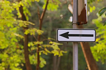 Left traffic route only direction sign turn pointer, background in park, black arrow icon on white frame road sign, grey pole post
