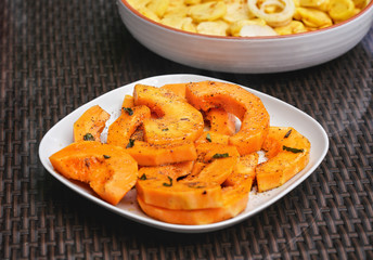 Grilled orange butternut squash pieces with herbs, on white square plate, another full of potatoes in background