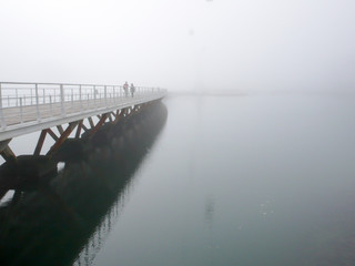 pedestrians crossing a long wooden bridge over calm water on a foggy and misty morning