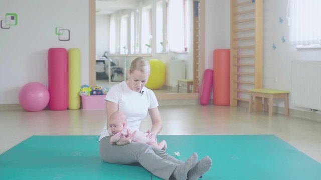 Woman swing little baby on legs back and forth. Wide shot of a physiotherapist gym with person in focus sit on floor.