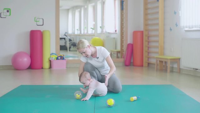 Physiotherapist playing with baby on foam in fast-motion. Wide shot of a physiotherapist gym with person in focus sit on floor.