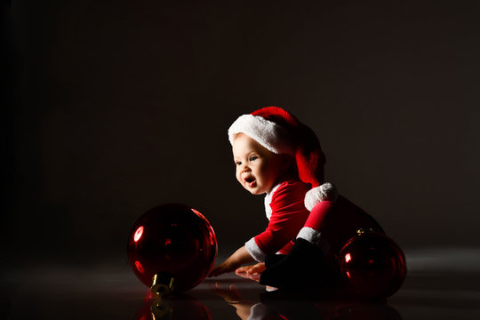 Amazed Infant Baby Boy Toddler In Red New Year Costume Is Playing With Christmas Tree Decoration, Rolling Fir Red Ball