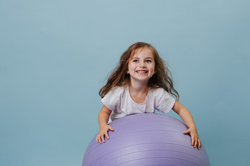 blue-eyed curly toddler girl laughs playing on lilac fitness ball.