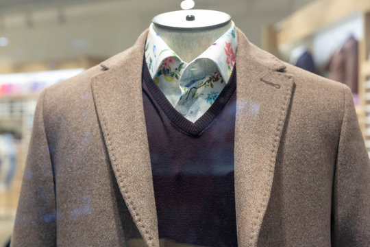 A beige coat, black, jumper and white shirt with a flower pattern hang on a mannequin behind glass in the window of a fashionable men's clothing store.