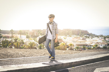 Trendy young boy using skateboard outdoor with bag on the shoulders. Happy teen come back from school with skate, exotic panorama in background. Freedom education carefree youth and lifestyle concept.