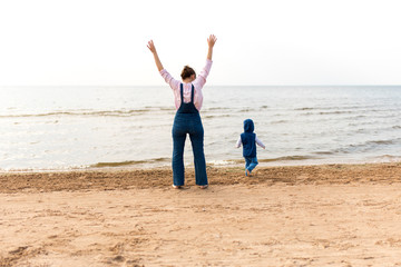 Woman with a child on the beach in front of open water. The child runs away.