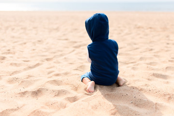 unrecognizable child sits with his back in a sweater with a hood on the beach with bare legs.