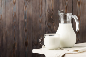 Glass jug and mug with milk on the edge of a white wooden on a background of brown wooden boards.