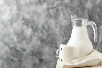 Glass jug and mug with milk on the edge of a white wooden on a concrete background.