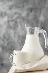 Glass jug and mug with milk on the edge of a white wooden on a concrete background.