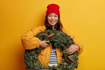 Pleasant looking woman with gentle smile holds green handmade wreath, wears hat and jacket, enjoys winter holidays, isolated over yellow wall, prepares Christmas gift. Xmas circlet with cones