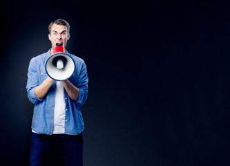 Excited man shouting through megaphone, with empty copy space area for slogan, advertising or text, against black background. Caucasian male model in blue clothing making announcement.