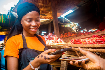 young african woman selling tomatoes in a local african market receiving payment via mobile phone transfer - obrazy, fototapety, plakaty