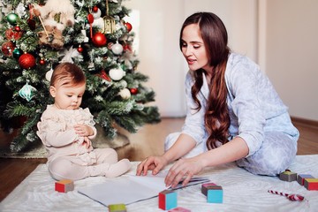 Happy beautiful woman having fun with her little cheerful daughter near the Christmas tree. Mom reads a book to the child and plays with colored cubes at home