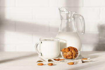 Glass jug and mug with almond milk on a white wooden table, lit by morning sunlight through the window. Bowl with almonds in the kitchen.