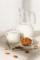 Glass jug and mug with almond milk on a white wooden table, lit by morning sunlight through the window. Bowl with almonds in the kitchen.