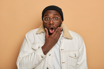 Portrait of shocked black hipster guy opens mouth, holds breath from surprisement, wears white stylish clothing, optical glasses, feels impressed and very excited, isolated on brown background