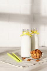 Two glass bottles with almond milk, decorated with a yellow ribbon on a white wooden table. Illuminated by morning sunlight through a window. Two cocktail rolls and a bowl of almonds in the kitchen.