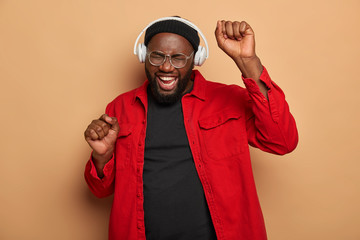 Happy joyful man with bristle raises hands and enjoys nice vibes of favourite song, listens music in headphones, wears red shirt, poses over brown studio wall, has energetic mood, feels amused