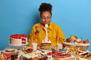 Ethnic sweet tooth holds spoon with cream, enjoys eating tasty dessert, poses at table with...