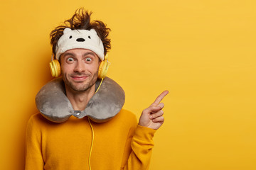 Happy European man points away with fore finger, listens favourite music via headphones, flies in airplane, wears blindfold and neck pillow, connected to some device, advertises blank space.