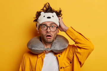 Impressed Caucasian man rubs head, keeps mouth opened, wears sleep mask and travel pillow, stares through spectacles, wears yellow shirt, travels on long distances. Rest in transport concept