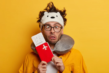 Funny male traveler crosses eyes, feels bored to wait long time for departure, wears transpaent glasses, sleep mask and neck pillow, stands with flying tickets and passport, isolated on yellow wall