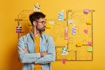 Horizontal shot of pensive bearded man stands with crossed arms, turns away, studies house design...