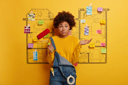 Hesitant curly woman stands with paint roller, thinks how to refurbish house, chooses new color for room, poses against house design sketch, dressed casully. New housing and renovation concept