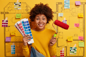 Smiling curly female with Afro hairstyle dressed in casual wear, holds color palette and paint...