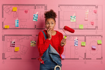 Pensive female homemaker applies red paint with roller, wears sweater and denim overalls, thinks...