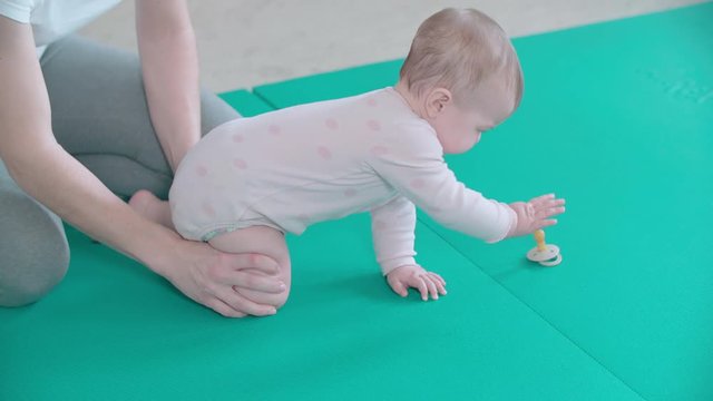 Physiotherapist force baby crawl on all fours in slow motion. Long shot slow motion of a baby in focus doing exercise on turquoise floor mat.