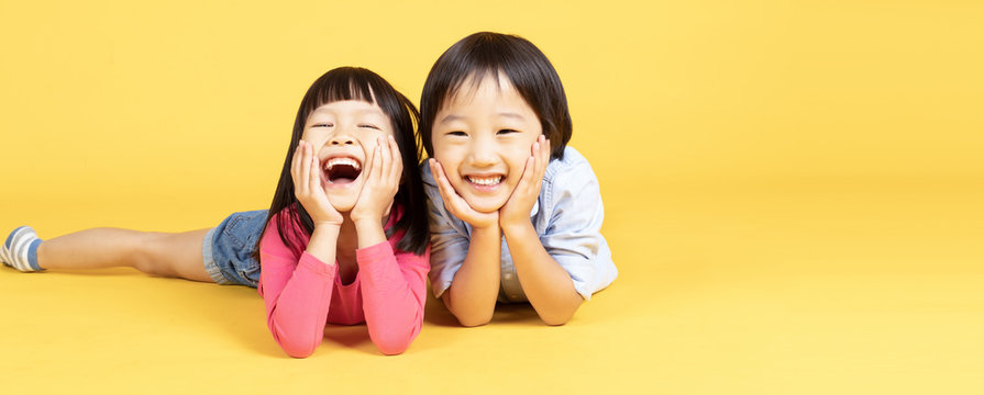Two Happy Asain kids lying on the floor and touches cheeks with both hands, has broad smile, shows perfect teeth over yellow lights banner  background. Banner panorama.