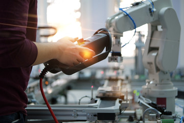 Close up of man's hand holding teach panel  to control robot arm which is integrated on smart factory production line. industry 4.0 automation line equipped with sensors and robot at background.