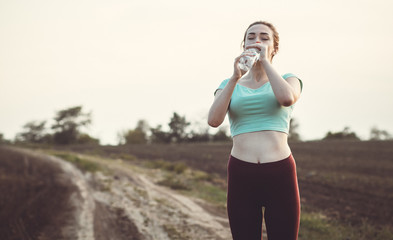fathletic girl drinking water after running a distance in a field at sunset, young woman quenches...