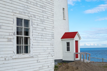 The exterior walls of two buildings. The white wood building in the foreground has paint peeling from the clapboard and a eight pane window. The second is the base of a lighthouse.