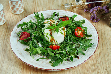 Tasty, fresh salad with mozzarella burrata, arugula, cherry tomatoes and basil on a gray plate on a wooden background. soft Italian cheese burrata. Italian cuisine. CLose up