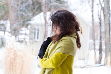 Medium horizontal portrait of cute brunette young woman standing outdoors in profile holding a miniature black rabbit cradled in her arms against a soft focus winter garden background