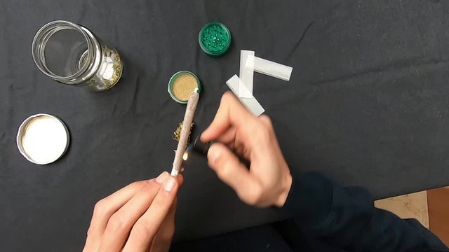 First person view of man rolling and smoking a joint of marijuana. Marijuana buds, grinder and crystal jar on black table. 