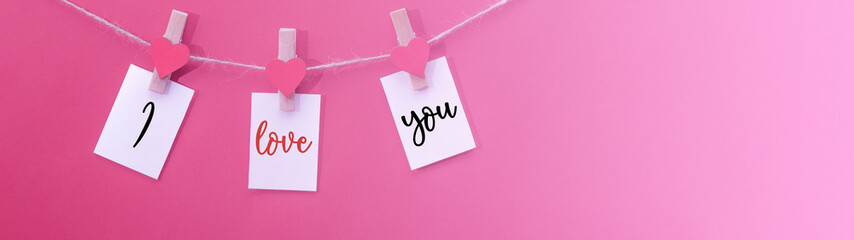 I love you - Clothes pegs with wooden hearts and paper notes hang on rope isolated on pink texture background panorama banner long, with space for text