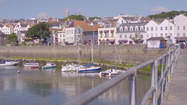 A handheld wde shot of a row of houses in a coastal town with boats moored and the edge of a bridge on a sunny day. Europe